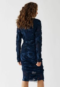Rochie bleumarin cu mâneci lungi, textură din material floral, design plisat și margine sub genunchi. Viziunea din spate arată detaliile modelului.