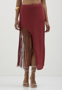 Jupe en tissu rouge avec une haute fente et un long détail en frange tressée d'un côté ; texture lisse et design longueur cheville.