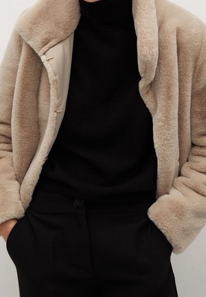 Osoba oblečená v béžovém kožichu z umělé kožešiny přes černý rolák a černé kalhoty, ruce v kapsách, zobrazená od krku po boky.