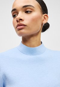 Φωτεινό γαλάζιο πουλόβερ με ψηλό λαιμό, κατασκευασμένο από υφασμάτινο πλεκτό με υφή. Συνοδεύεται από μικρά ασημένια κρίκους σκουλαρίκια.