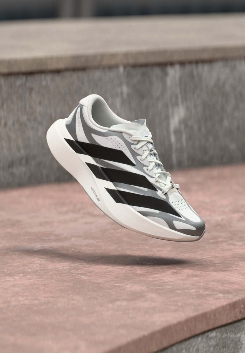 Chaussure de course blanche et grise avec des rayures noires, flottant au-dessus d'une surface en béton rosée près d'un mur en pierre grise.