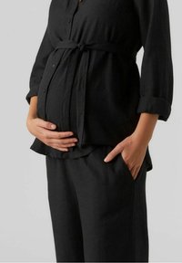 Czarna sukienka ciążowa z koszulą zapinaną na guziki przewiązaną w talii oraz luźnymi spodniami; miękka tkanina i luźny krój podkreślają komfort.