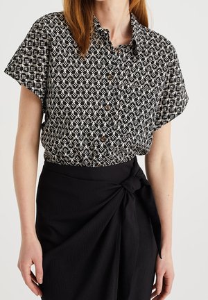 Kobieta w czarno-białej, krótkorękawowej bluzce na guziki z wzorem, wpuszczonej do czarnej spódnicy z ozdobnym wiązaniem z boku.