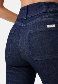 Jeans in denim blu scuro con vita alta, elastico in vita, due tasche posteriori e una piccola etichetta con la scritta "ITEM m6". Texture liscia.