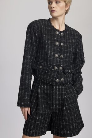 Giacca in tweed nera con motivo a quadri bianchi, dettagli in frange e bottoni argentati. Abbinata a pantaloni corti in tweed coordinati, con pieghe.