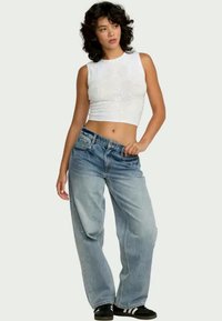 Junge Frau in einem weißen, ärmellosen Crop-Top, lockeren blauen Jeans und schwarzen Turnschuhen, steht mit einer Hand in ihrer Tasche.
