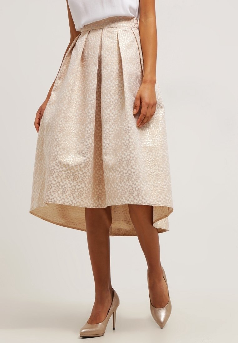 Jupe plissée couleur champagne clair avec un ourlet asymétrique, en tissu texturé présentant un motif subtil. Portée avec des talons beiges.