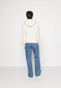 Una persona sta in piedi di spalle, indossando un felpa beige chiaro con il cappuccio abbassato e jeans blu, davanti a uno sfondo bianco semplice.