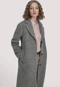 Frau mit lockigem Haar, die einen grauen langen Mantel über ein pink-weiß gestreiftes Hemd und eine beige Hose trägt, posiert mit einer Hand in der Manteltasche.