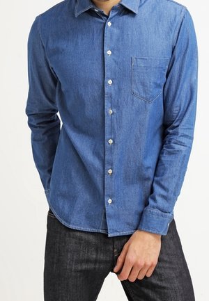 Mężczyzna ubrany w dopasowaną niebieską dżinsową koszulę na guziki z kieszenią na piersi oraz czarne dżinsy na białym tle.