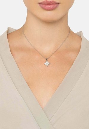 Hopeaketju, jossa on kukkaketju ja neljä pyöreää, valkoista kiveä. Ketju on ohut ja herkkä, ja se on asetettu vaaleanbeigeä toppia vasten.