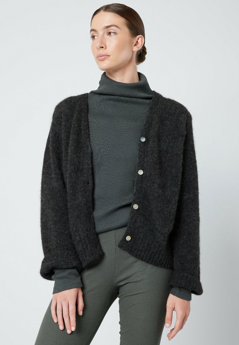 Temno siva volnena jopica z ohlapnim krojem, z rebrastimi manšetami, z gumbom za zapiranje in mehko teksturo, preplastena čez temno zeleno turtleneck.
