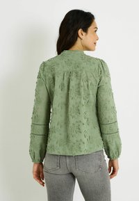 Blusa verde bordada con motivos florales, tela texturizada, mangas largas con detalles de encaje y escote redondo.