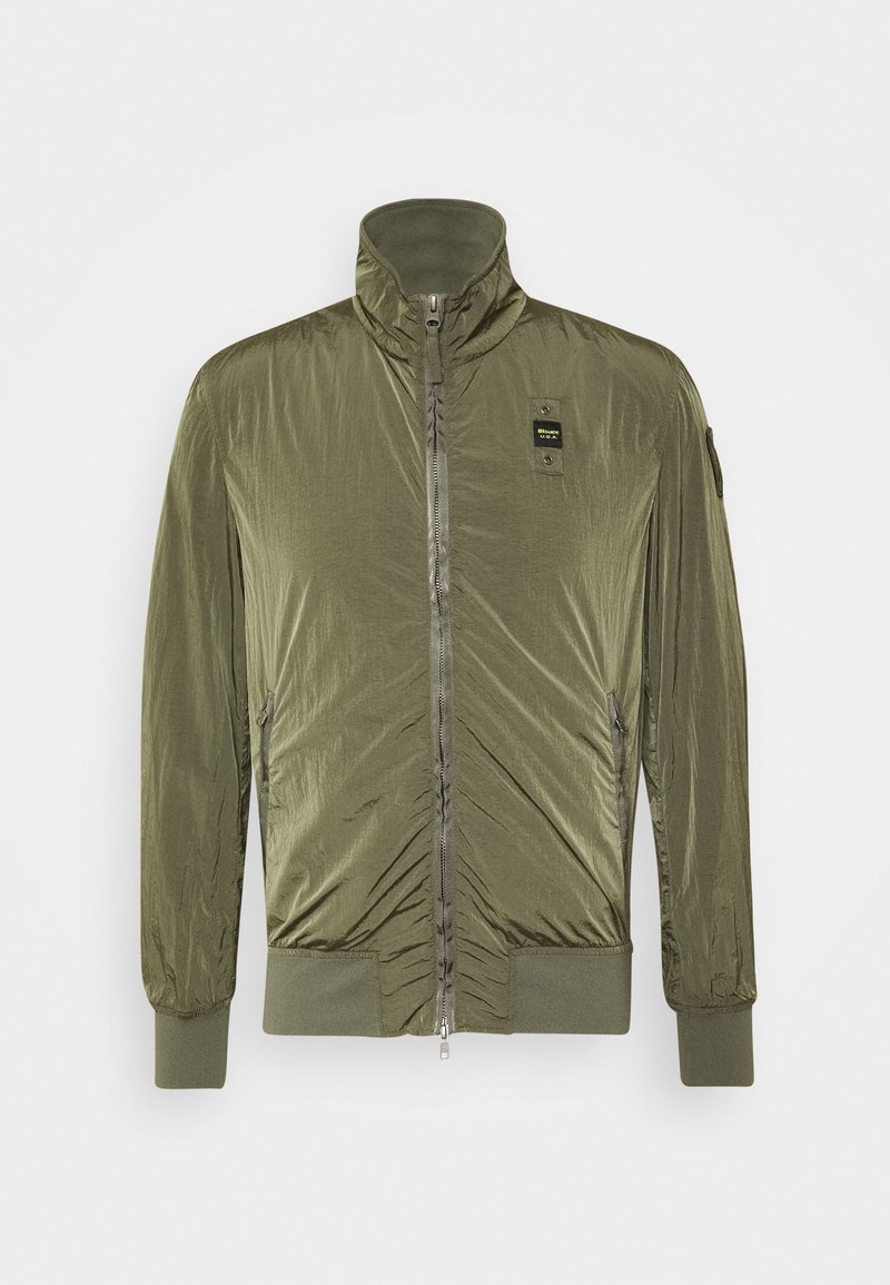 Veste légère zippée vert olive avec col montant, poignets et ourlet côtelés, et un petit patch logo noir sur la poitrine.