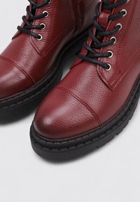 Botines de cuero texturizado rojo con cordones y ojales negros. Punta redonda y suelas gruesas de goma negra con detalles de costura.