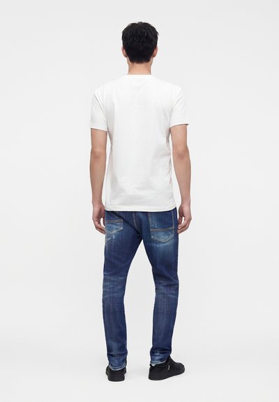 Homme debout de dos, portant un t-shirt blanc uni à manches courtes, un jean bleu avec des poches et des baskets noires sur fond blanc.