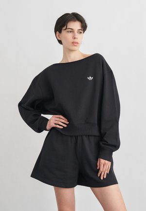 Ung person med kort mørkt hår, iført løstsiddende sort sweatshirt med hvid Adidas-logo og matchende sorte shorts, poserer foran en grå baggrund.