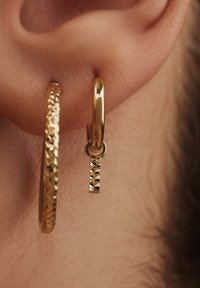 Gold hoop and pendant earrings featuring a textured surface and a small square-patterned charm dangling from the hoop.