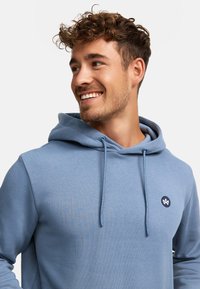 Lyseblå hettejakke laget av mykt stoff med snøring i hetten. Har en rund logo i mørkere blå på brystet. Glatt tekstur, enkel design.