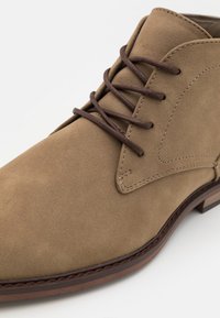 Brune suede ankelstøvle med brune snørebånd, rund tå og subtile syninger. Gummisål og glat tekstureret overflade.