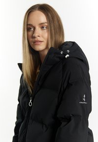 Jachetă puffer neagră cu fermoar frontal, având o glugă și șnur de ajustare. Mânecă stângă cu un logo alb în formă de ancora. Țesătură moale, texturată.
