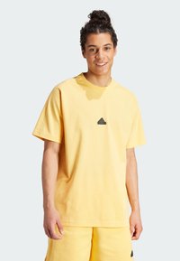 Żółta bawełniana koszulka z okrągłym dekoltem i krótkimi rękawami, z małym czarnym logo Adidas umieszczonym na środku klatki piersiowej. Gładka tekstura.