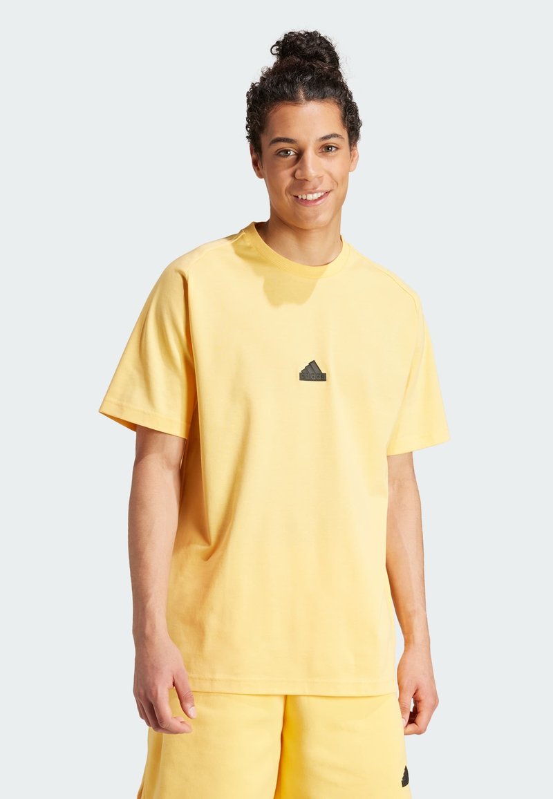 Żółta bawełniana koszulka z okrągłym dekoltem i krótkimi rękawami, z małym czarnym logo Adidas umieszczonym na środku klatki piersiowej. Gładka tekstura.