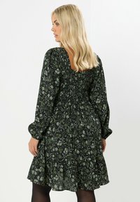 Musta mekko, jossa on vihreä kukkakuosi, pitkät puffi-hihat, smokkattu vartalo ja röyhelöhelma. Vaate virtaa kankaasta, jolla on teksturoitu ulkonäkö.