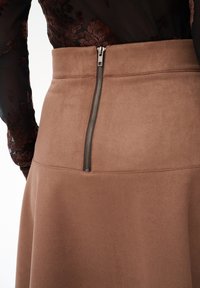 Zbliżenie brązowej spódnicy z zamszu z widocznym zamkiem błyskawicznym z tyłu, noszonej z przezroczystą czarną bluzką w kwiatowy wzór.