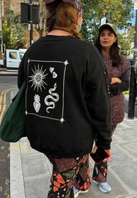 Čierna mikina s bielym grafickým motívom slnka, hada, ananásu, sŕdc a dekoratívnych okrajov. Kombinovaná s nohavicami s potlačou.