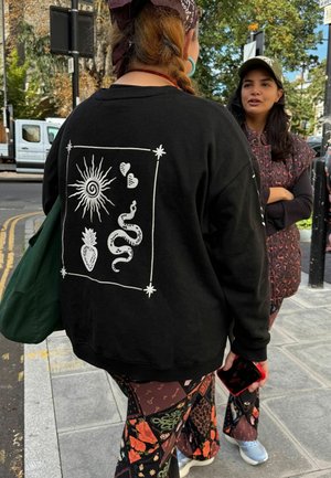 Melnās krāsas džemperis ar baltu grafisku dizainu, kurā attēlots saule, čūska, ananāss, sirdis un dekoratīvas apmales. Kombinēts ar rakstainām biksēm.