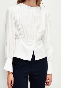 Witte blouse met lange mouwen, voorzien van een subtiel patroon, met een geplooide taille, ronde halslijn en uitlopende mouwen, gecombineerd met donkere, op maat gemaakte broek.