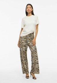 Beige t-shirt med präglad text, kombinerat med vida byxor med ett svart och tan randigt mönster som föreställer tigerstripor och svarta klacksandaler.