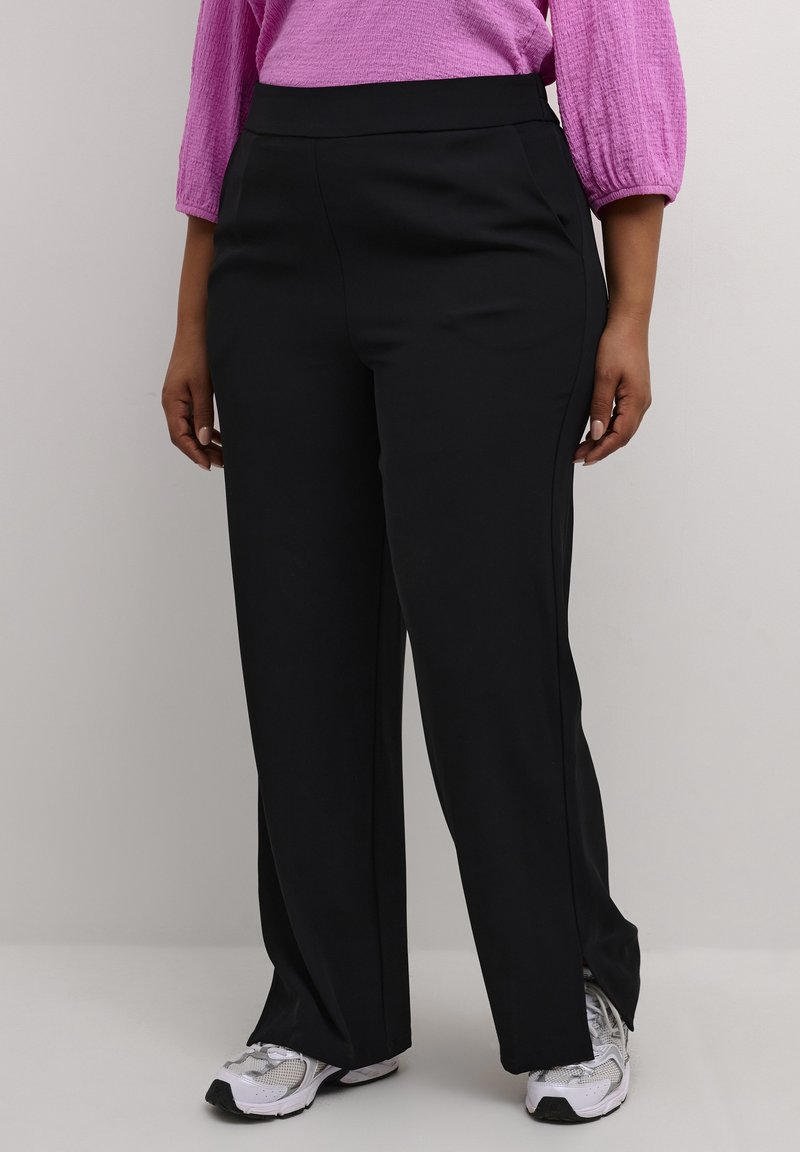 Personne portant un pantalon noir à jambes larges avec des fentes sur les côtés, des baskets blanches, et un haut rose à manches longues texturé, debout devant un fond gris.