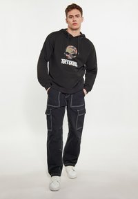 Fekete kapucnis pulóver "TUFFSKULL" felirattal, álcázott logóval. Fekete cargo nadrággal és fehér sportcipőkkel párosítva, eltérő varrással.