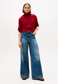 Suéter de punto trenzado rojo con cuello alto, combinado con jeans azules de pierna ancha y cintura alta. Sandalias de cuero marrón. Fondo simple.