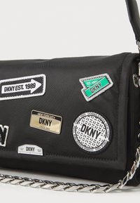 Black nylon handbag featuring multiple patches and metallic accents, including a chain strap and brand logo details.