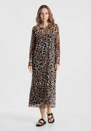 Kobieta stojąca, ubrana w długą suknię w panterkę sięgającą kostek oraz czarne sandały na otwartym palcu z klamrami.