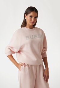 Roza dukserica s okruglim izrezom i prevelikim rukavima. Sadrži sjajni "GUESS" logo na prednjoj strani. Uparena s odgovarajućim hlačama.