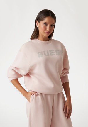 Sudadera rosa con cuello redondo y mangas anchas. Presenta un logo brillante de "GUESS" en la parte delantera. Combinada con un pantalón a juego.