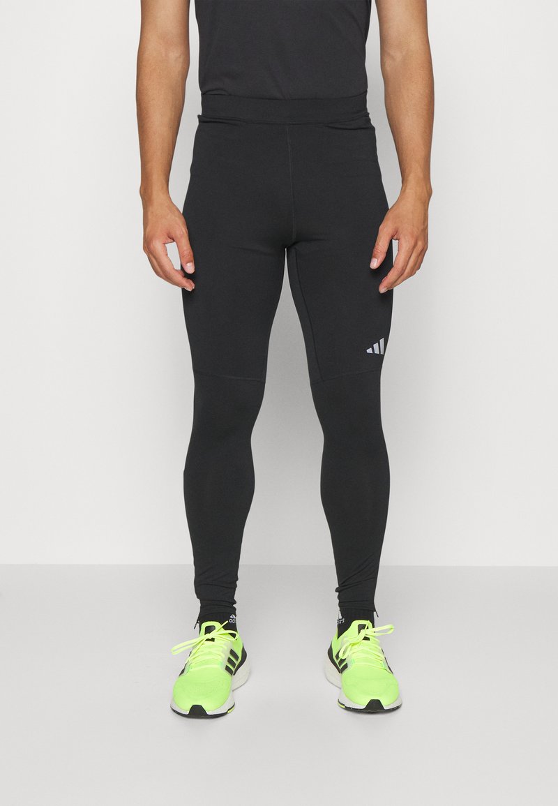 Zwarte sportleggings met een aansluitend ontwerp, gemaakt van rekbare stof, met een logo aan de linkerzijde en gedragen met neongroene sneakers.