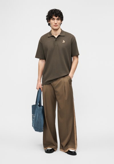 Νεαρός με σγουρά μαλλιά που φοράει σκούρο καφέ πόλο, φαρδιά καφέ παντελόνια, μαύρα παπούτσια, κρατώντας μια μπλε τσάντα tote, στέκεται σε λευκό φόντο.