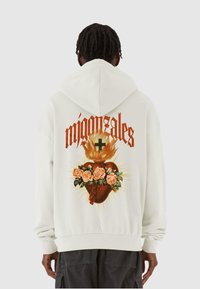 Hvid hoodie med et stort grafisk design på bagsiden. Den inkluderer et brændende hjerte, roser og ordet "mijonzales" i fed orange skrift.