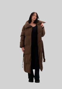 Giacca a vento marrone con colletto alto, caratterizzata da una silhouette lunga e grandi pannelli trapuntati. Include una cintura staccabile per la chiusura.