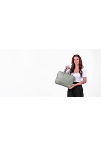 Grüne Lederhandtasche mit zwei Griffen, einem gesteppten Muster und einem Reißverschluss, gehalten von einer Frau in einem weißen Oberteil und dunklen Hosen.