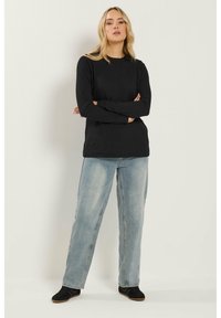 Blondynka stojąca z założonymi rękami, ubrana w czarną bluzkę z długim rękawem, wyblakłe niebieskie dżinsy i czarne trampki na białym tle.