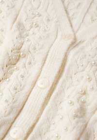 Kremowy sweter z dzianiny z perłowymi koralikami naszytymi w wzór warkoczowy oraz lekkimi, przezroczystymi guzikami na całej długości przodu.