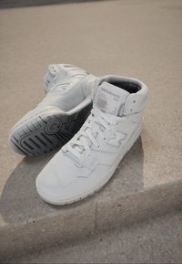 Białe trampki wysokie wykonane ze skóry, z perforowanymi detalami, płaskimi sznurowadłami, wyściełanym kołnierzem oraz szarym gumowym spodem z wzorem zapewniającym przyczepność.