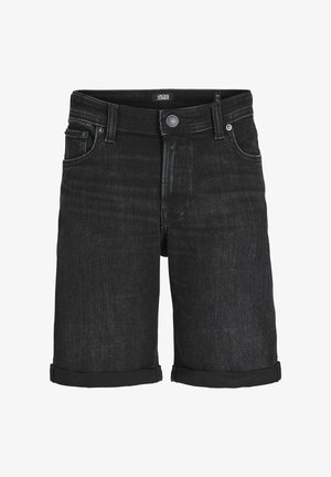 Person iført en grå hættetrøje med teksten "JACK & JONES", sorte opkrøllede denimbukser, sorte sokker og hvide sneakers, stående mod en ensfarvet baggrund.