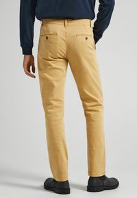 Pantalon chino kaki à texture lisse, coupe slim, avec deux poches arrière. Porté avec des bottines noires, vu de dos.